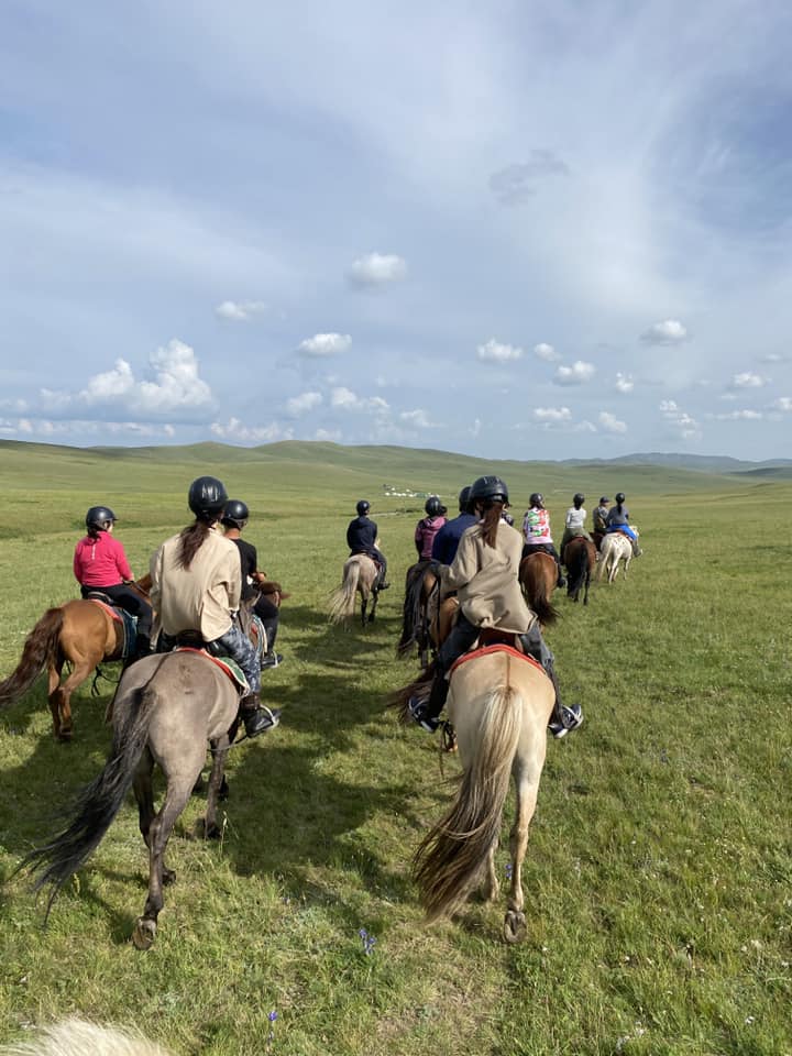 Das Bild zeigt eine Gruppe von Reiterinnen und Reitern in der Mongolei. Grüne Wiese, leicht bewölkter Himmel