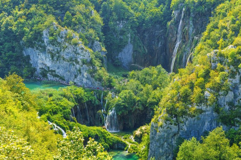 Das Bild zeigt die Berge, davor 2 türkisfarbene Seen, die Kaskadenförmig ineinander übergehen. Die Berge sind mit grünem Moos bedeckt. Nationalpark Plitvicer Seen
